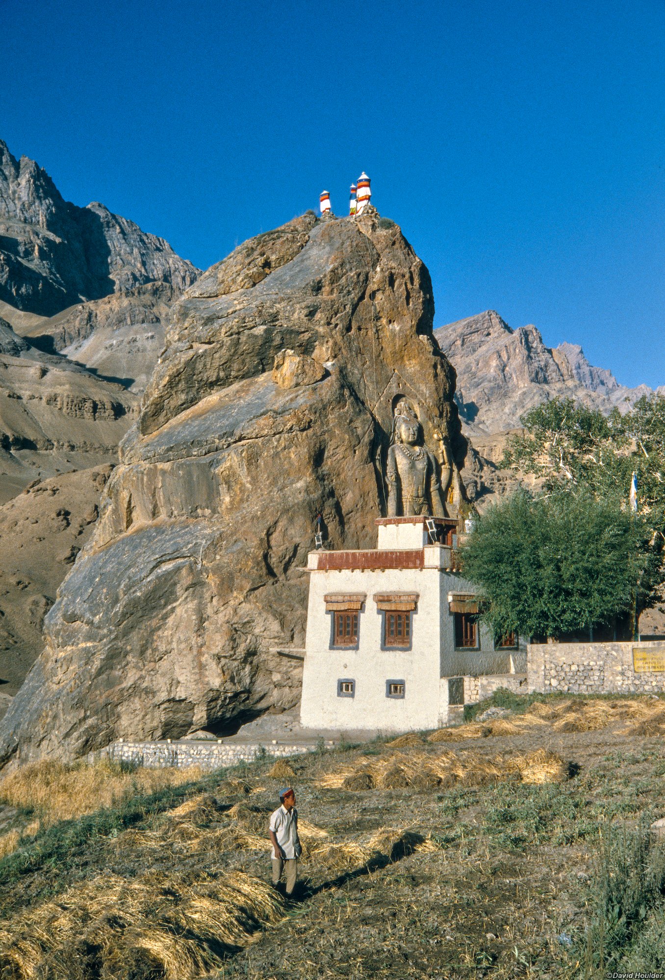 A large Buddha figure carned into a rockface with a small building at its base, and a man harvesting a crop in the foreground