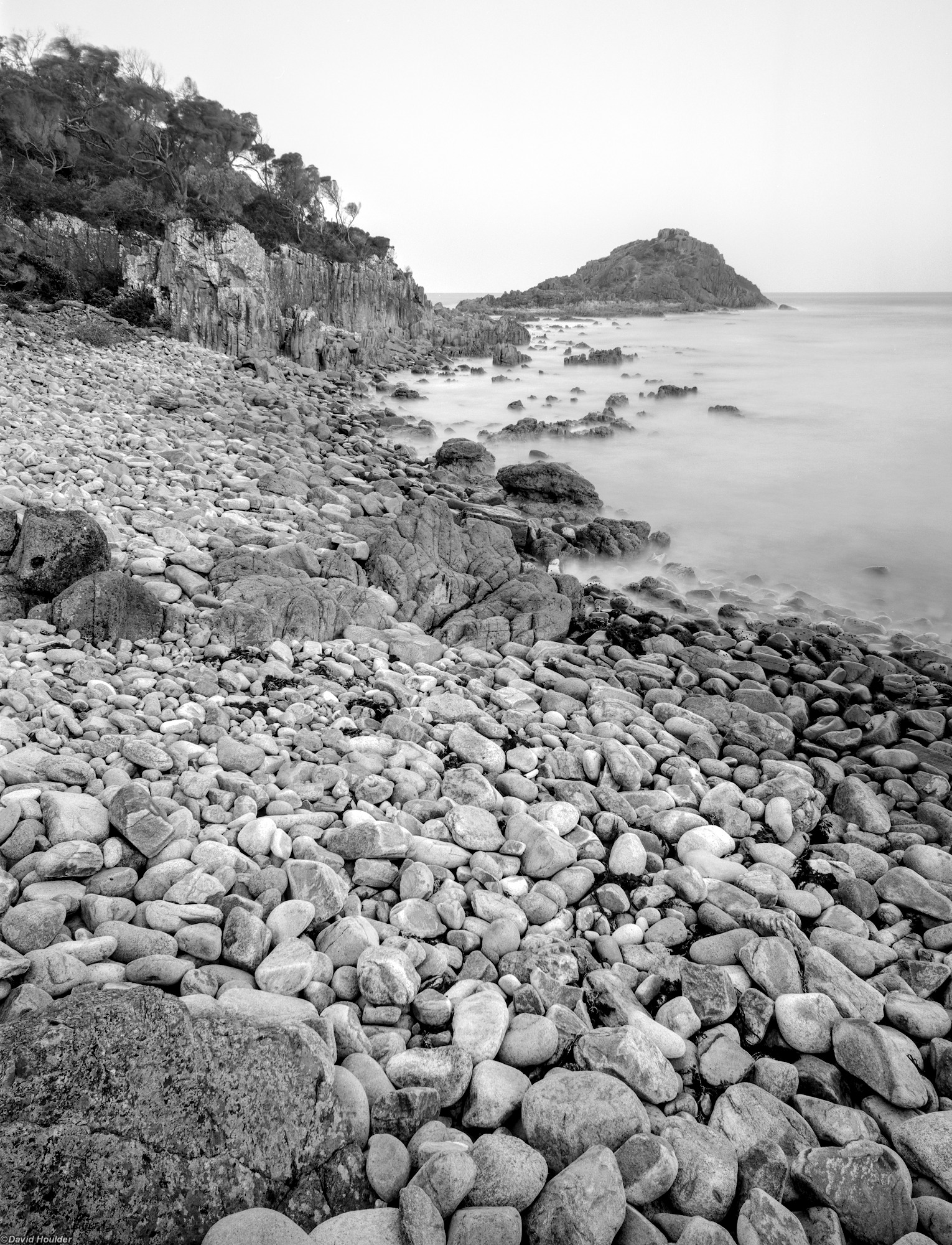 An ocean bay with a headland and a shore of rounded rocks in the foreground