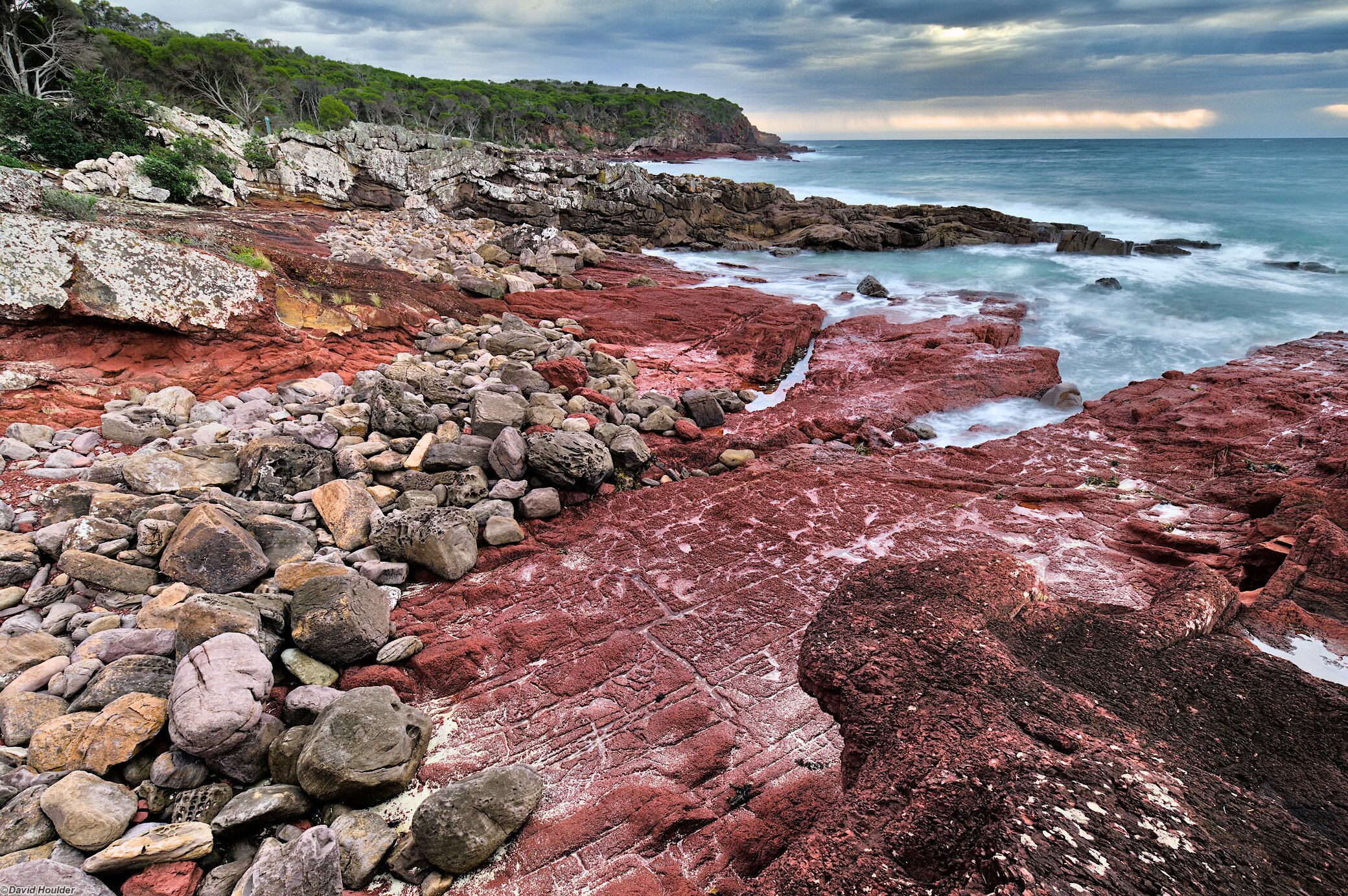 Ocean shore with boulders sitting on a rock shelf and a forested headland in the distance