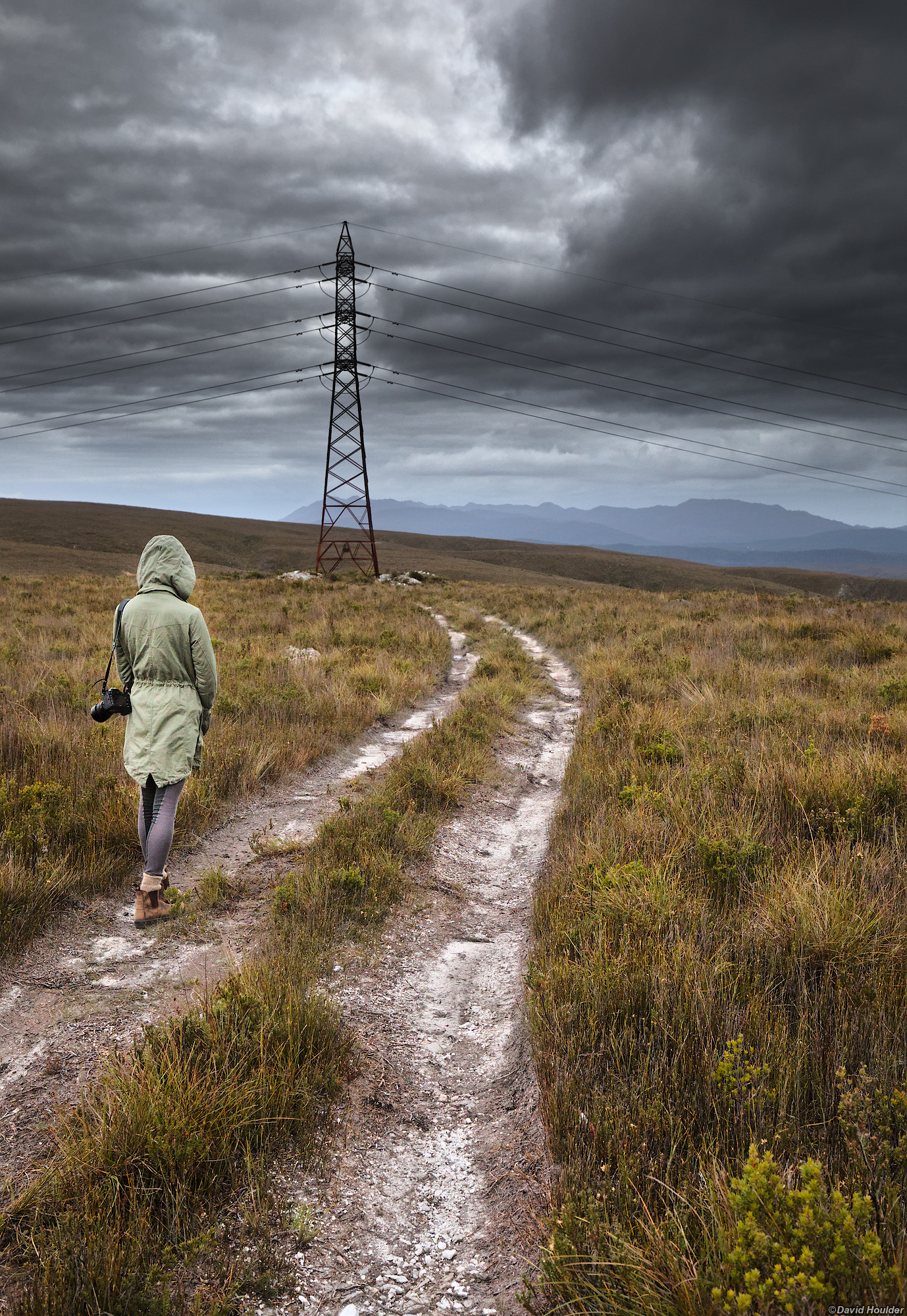 A young woman in a rainjacket walking away from the camera along a track through alpine grassland towards a large electricity pylon.
