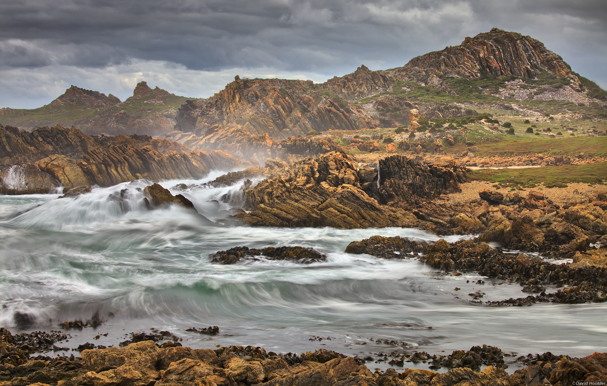 Ocean waves crashing onto rocks with dark clouds and rocky outcrops behind