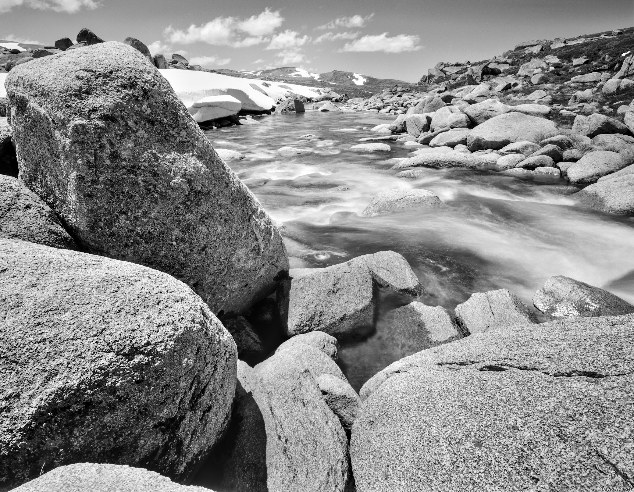 Large granite boulders beside  a flowing river and patches of snow in the distance