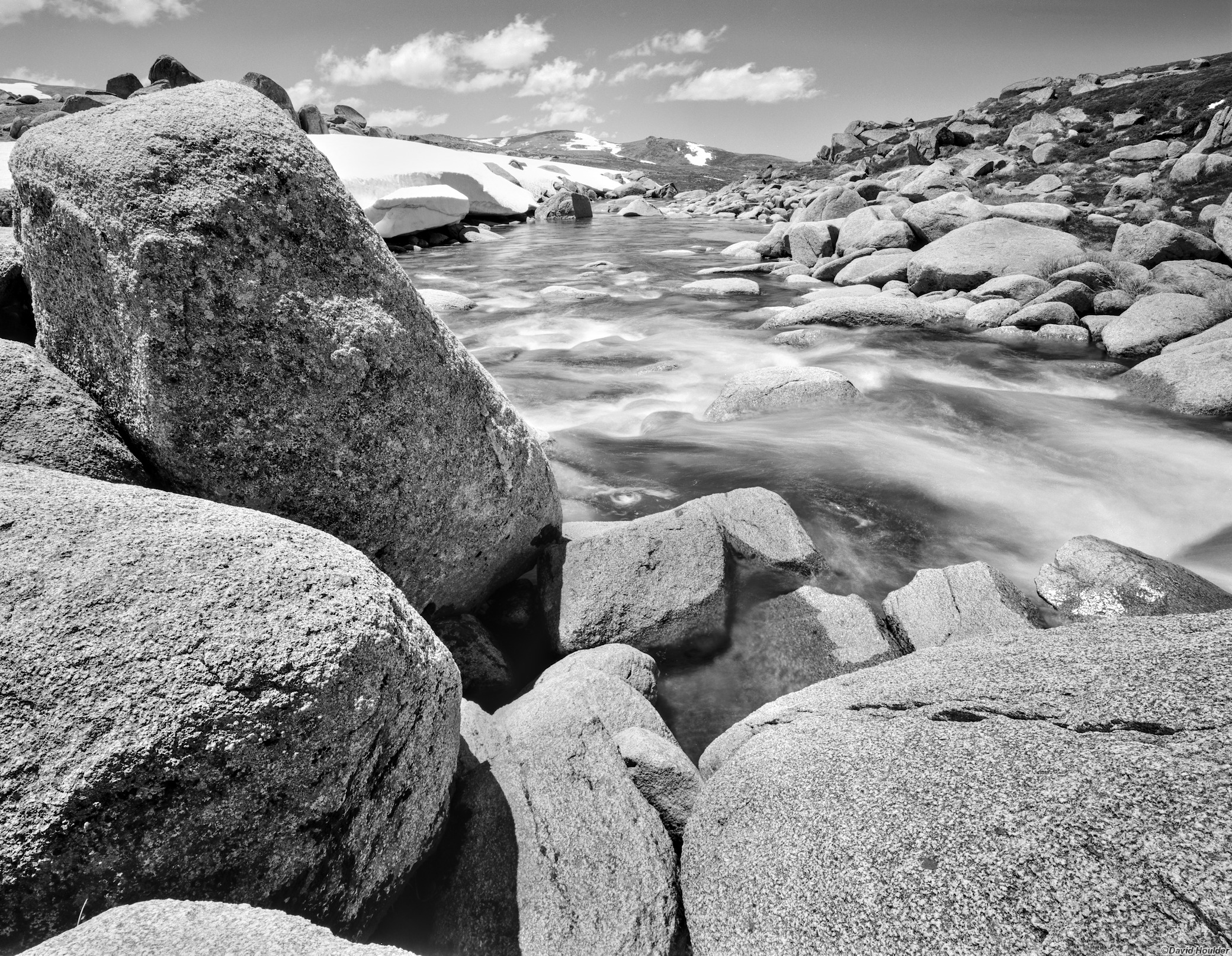 Large granite boulders beside  a flowing river and patches of snow in the distance